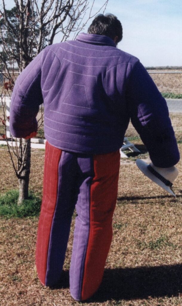 Person wearing oversized, padded purple jacket and pants, holding a helmet, standing outdoors near a leafless tree.