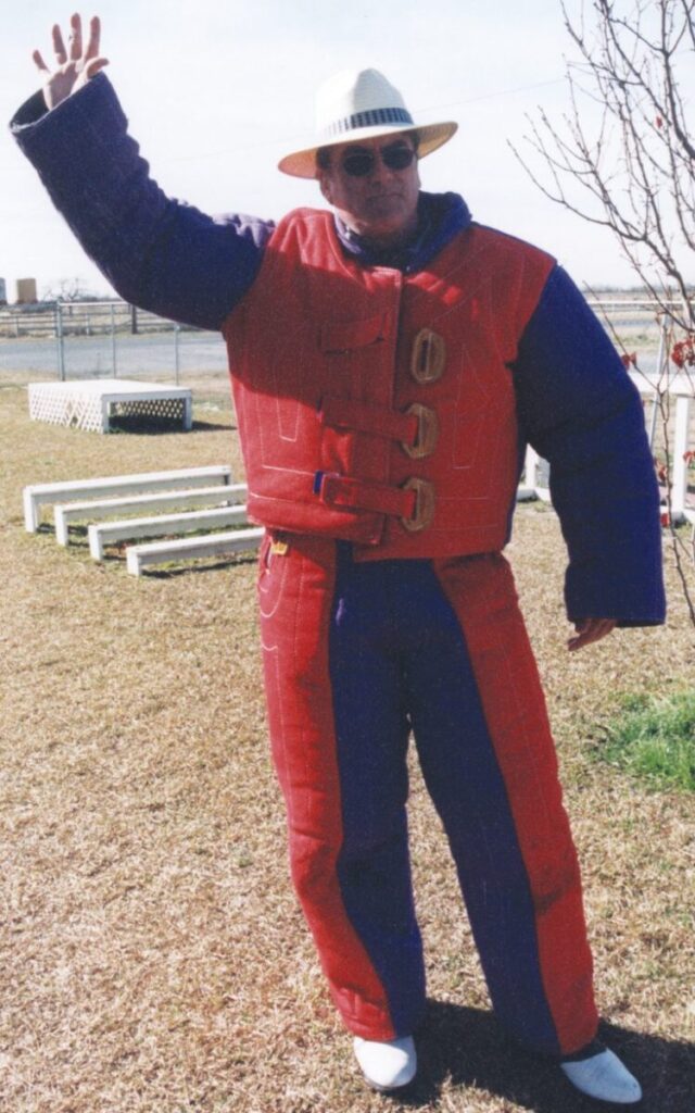 Person in a padded red and blue suit waves with one hand, wearing a hat and sunglasses, standing on grass near a tree.